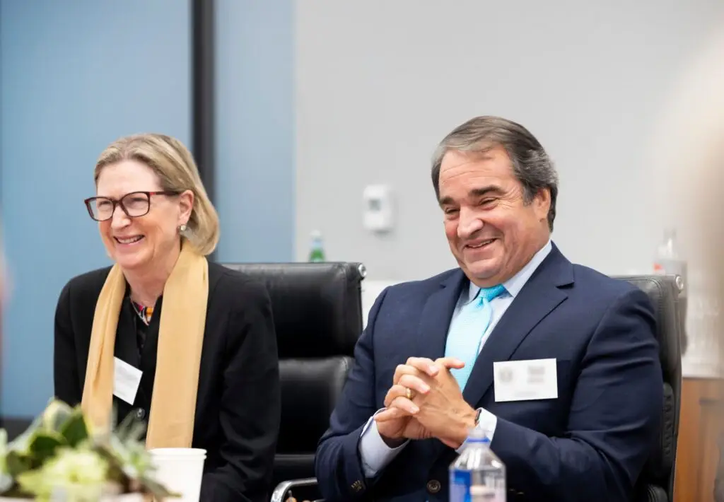 Woman and man in a conference room. Both people are smiling. Woman is on the left with short blonde hair and she is wearing glasses, yellow scarf, and a black blazer. Man is wearing a suit.